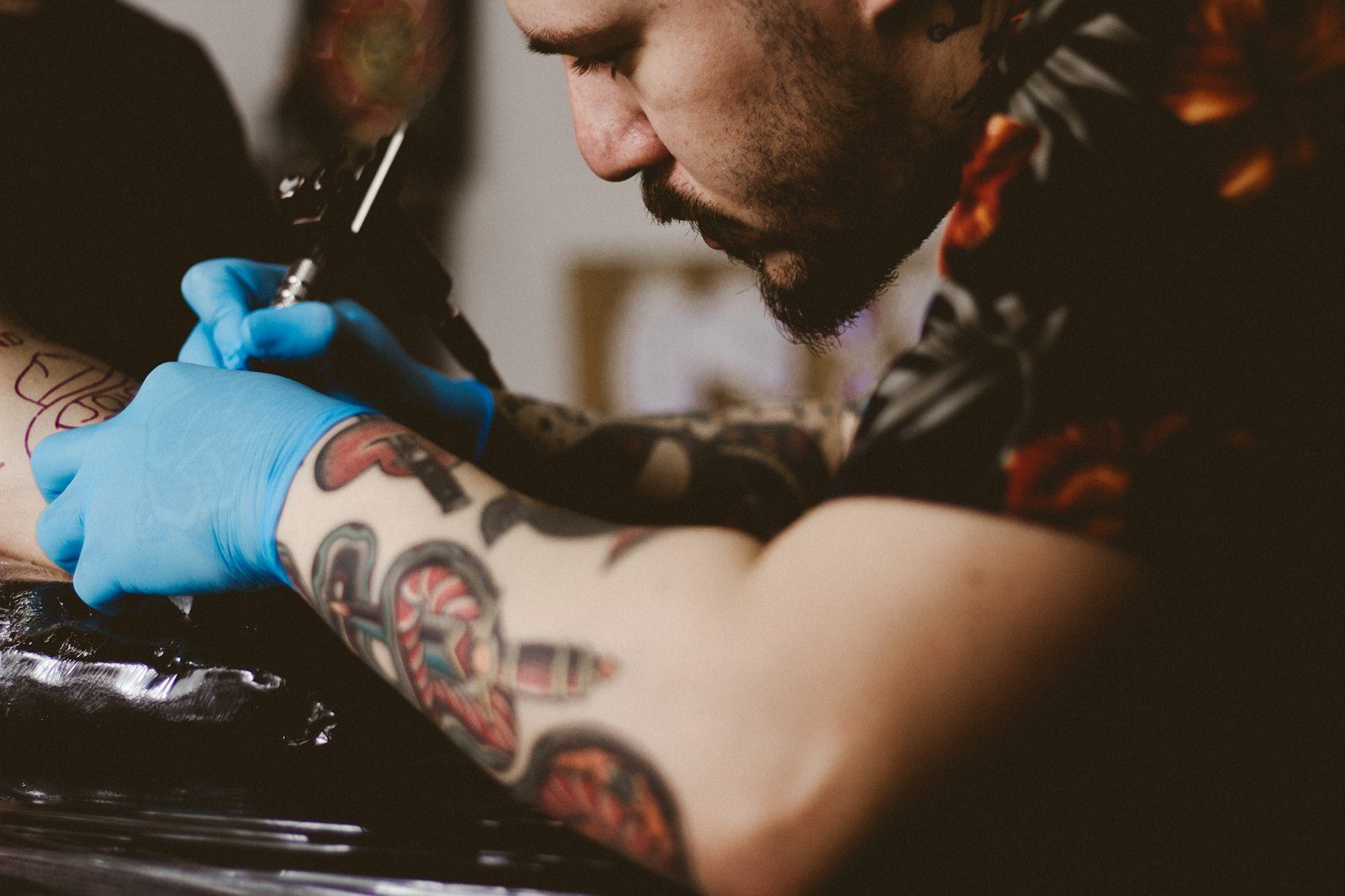Moderne Website für ein Tattoostudio, Beispielansicht: Tätowierer mit Bart und blauen Handschuhen setzt die Nadel am Unterarm eines Kunden an, seine eigenen Arme sind farbig tätowiert